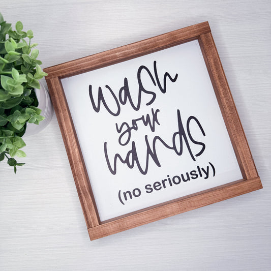 Framed sign with humorous text 'Wash your hands (no seriously)' on a light background