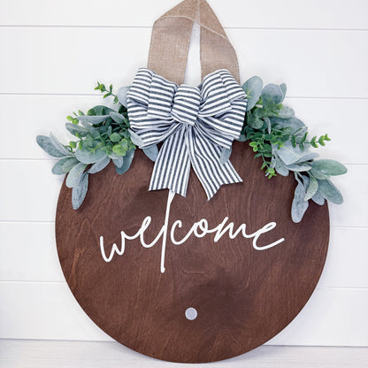 Wooden door hanger with 'welcome' text, striped bow, and greenery on a white background