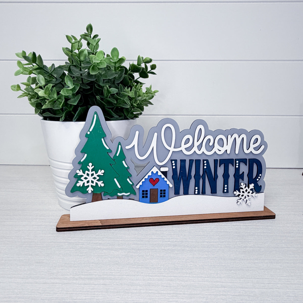 A decorative wooden tabletop sign reading 'Welcome Winter' with a green tree, blue house, snowflakes, and a small white snowdrift, displayed on a light gray surface with a potted green plant behind it.