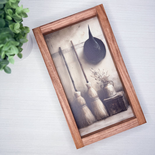A rectangular wooden sign containing a vintage-style illustration of three broomsticks, a black hat, and a small vase, displayed on a light gray wood-textured surface next to a green plant.
