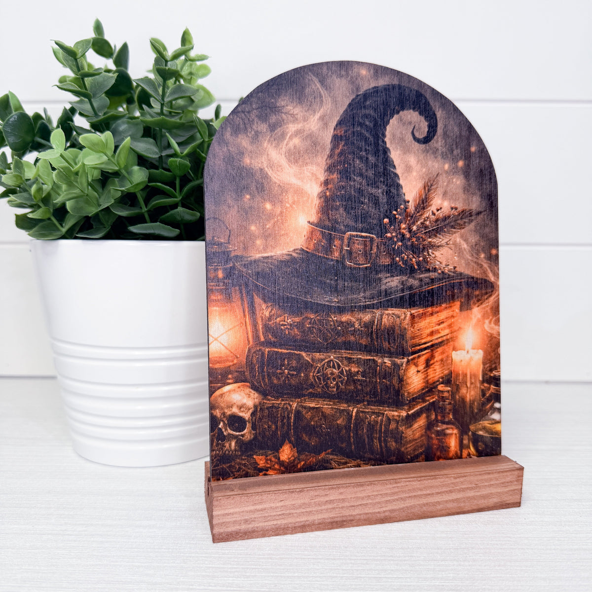 A decorative wooden plaque with an arched top and a small wooden base, featuring a printed witch hat resting on old books with candles, a skull, and autumn leaves. The plaque is styled on a white surface next to a potted green plant.