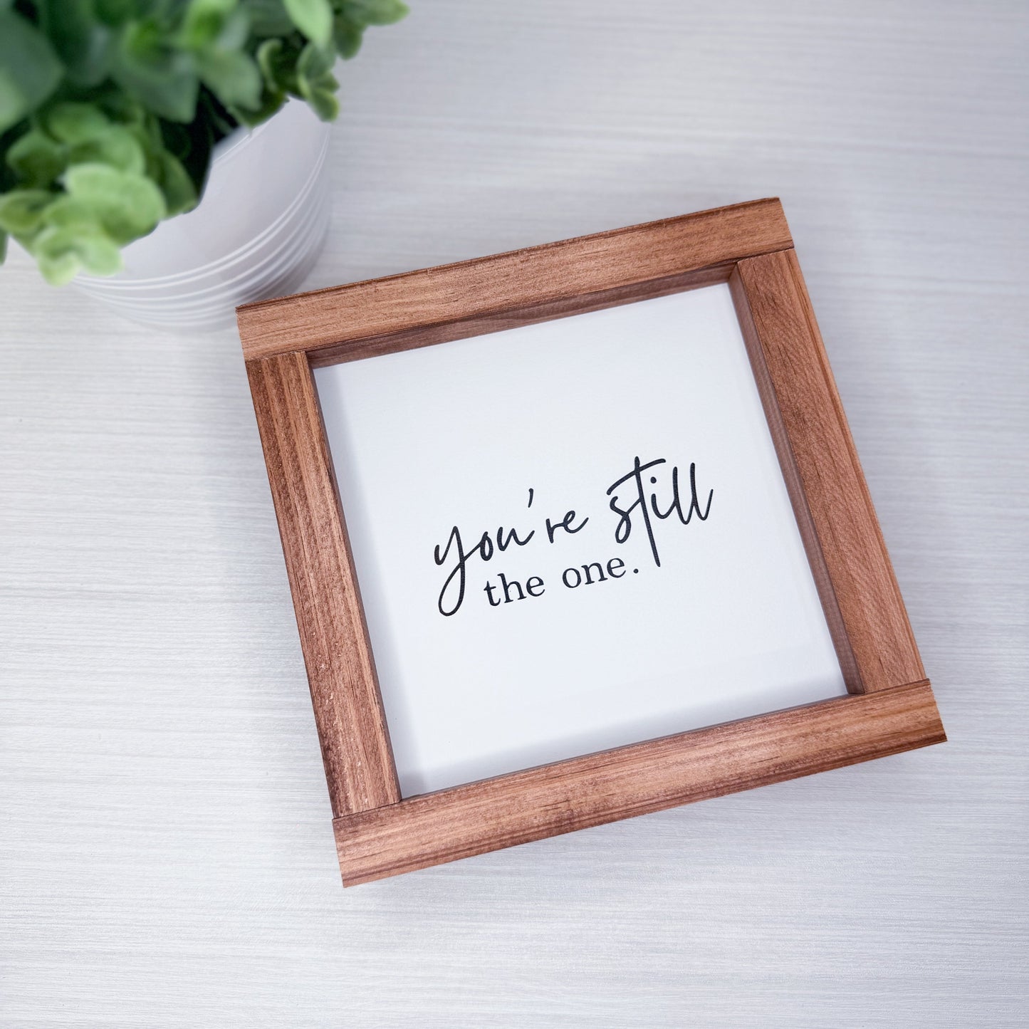 Wooden picture frame with a white card inside displaying 'you're still the one.' text, placed on a light surface with a plant in the background.